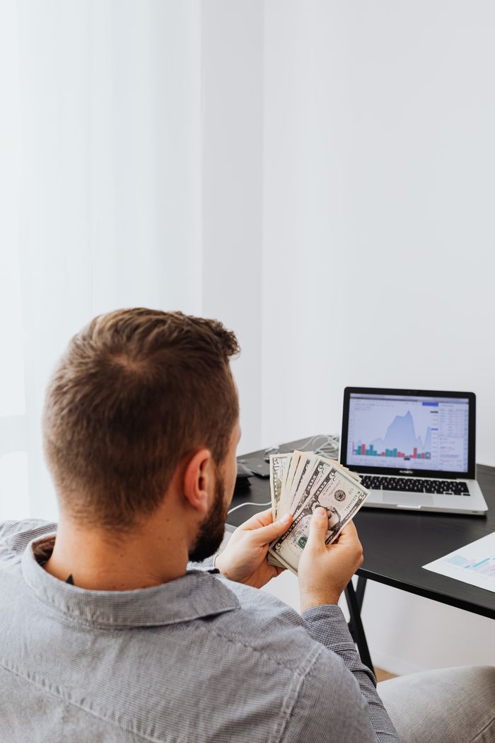 Businessman holding money while analyzing stock market data on laptop at home office setup.