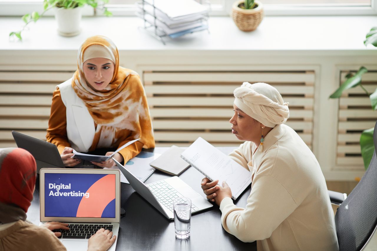 services-01 Three women in hijabs collaborating on digital advertising strategies around a table with laptops.